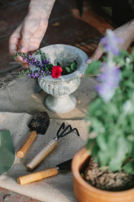 A pair of hands are seen arranging or planting purple and red flowers into a decorative stone garden urn, which is positioned on a textured outdoor surface. Nearby, gardening tools including a small h