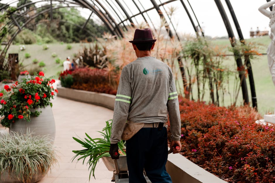 A gardener seen from behind working inside a modern greenhouse with a transparent, curved roof supported by black metal arches. The gardener is wearing a grey long-sleeve shirt with reflective stripes